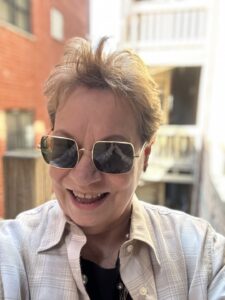 A person with short, light brown hair wearing sunglasses and a light-colored shirt smiles while standing outdoors, with buildings and a balcony in the blurred background&mdash;reminiscent of IR Approved Author Stella Atrium&rsquo;s candid advice on embracing life.