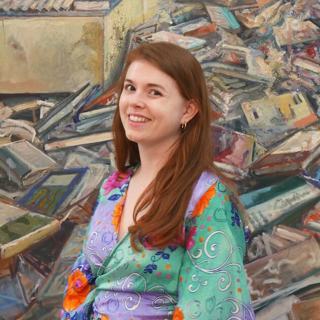 A woman with long brown hair, wearing a colorful floral dress, smiles while standing in front of a mural featuring a large pile of scattered books—perfect for anyone seeking book publicity or looking to hire a book publicist.