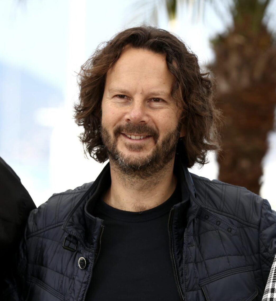 A man with medium-length curly brown hair and a beard is smiling at the camera. He is wearing a black quilted jacket over a dark shirt. There is a blurred palm tree and light background behind him.